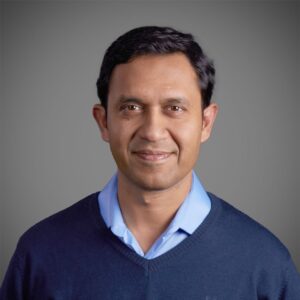 Retrato de homem com expressão leve sorridente, usando camisa azul clara e suéter azul escuro, em fundo neutro cinza