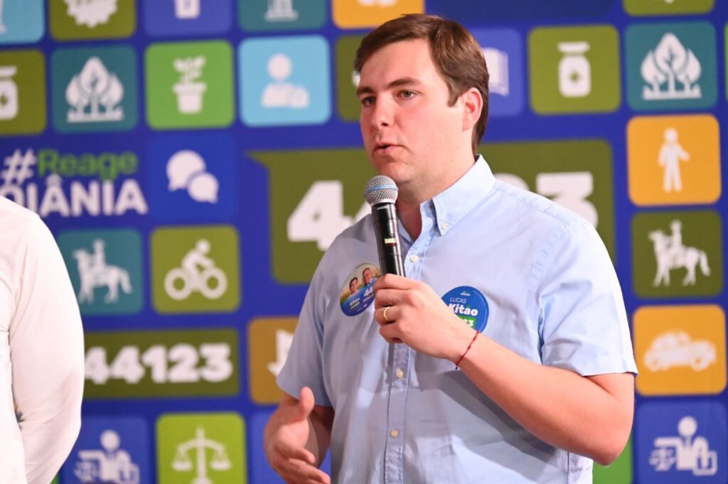 Homem falando ao microfone durante evento político, com painel colorido ao fundo.