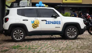 Carro branco com adesivo de abelha e logotipo Zenbee estacionado em frente a estabelecimento comercial com balões no teto