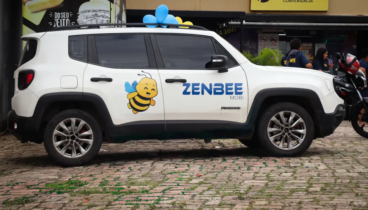 Carro branco com adesivo de abelha e logotipo Zenbee estacionado em frente a estabelecimento comercial com balões no teto