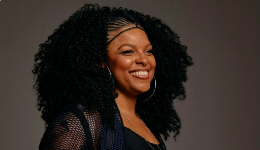 Mulher sorrindo com cabelo afro volumoso e tranças frontais, usando argolas e roupa preta.
