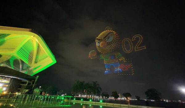 Show de drones formando personagem iluminado e número 02 no céu noturno sobre área à beira d’água