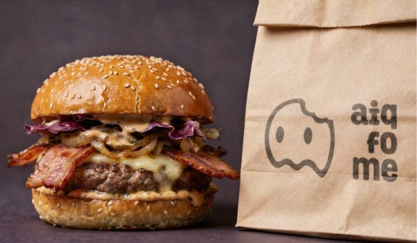 Hambúrguer artesanal com bacon, queijo derretido e pão com gergelim ao lado de sacola de papel da marca Aiqfome