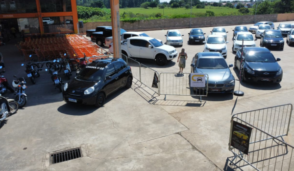Pátio de concessionária com diversos carros estacionados e área cercada por grades metálicas durante o dia.