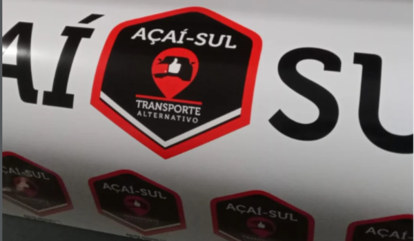 Adesivo da marca Açaí-Sul com a frase “Transporte Alternativo” aplicado na lateral de um veículo branco. O logotipo tem formato hexagonal, nas cores preto e vermelho, com um ícone de localização e um símbolo de “curtir” no centro.