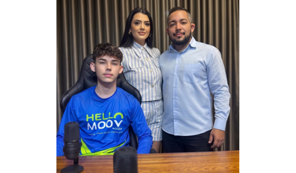 Três pessoas posam em um estúdio de podcast. Um jovem sentado em uma cadeira gamer usa camiseta azul da Hello Moov, enquanto um homem e uma mulher estão em pé atrás dele. À frente, aparecem dois microfones sobre uma mesa de madeira, com cortinas escuras ao fundo.