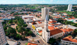Vista aérea de uma cidade com igreja de torre branca no centro, cercada por ruas, árvores e prédios residenciais.