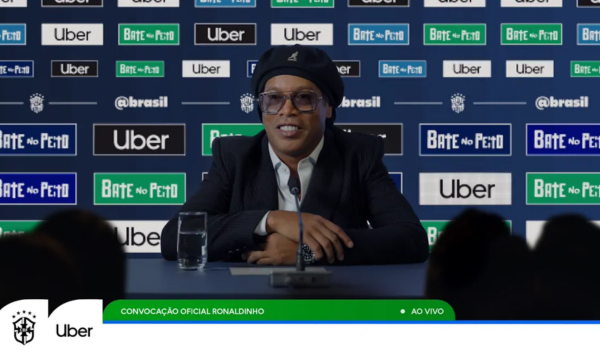 Ronaldinho Gaúcho participa de uma coletiva de imprensa em um evento patrocinado pela Uber e Bate no Peito. Ele está sentado diante de um microfone, usando terno escuro, boina preta e óculos, com painéis de marcas ao fundo.
