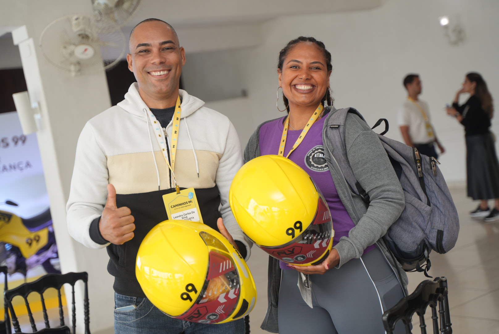 Dois motociclistas parceiros da 99 sorriem segurando capacetes amarelos durante evento Caminhos 99 no Rio de Janeiro.