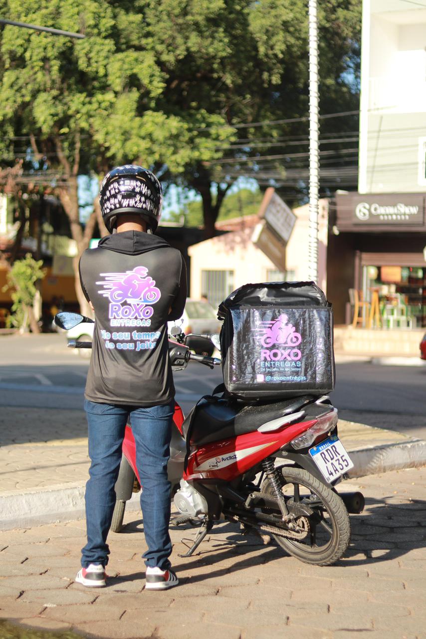 Entregador da Roxo Entregas ao lado de motocicleta com bag personalizada estacionada em rua de Barreiras, Bahia.