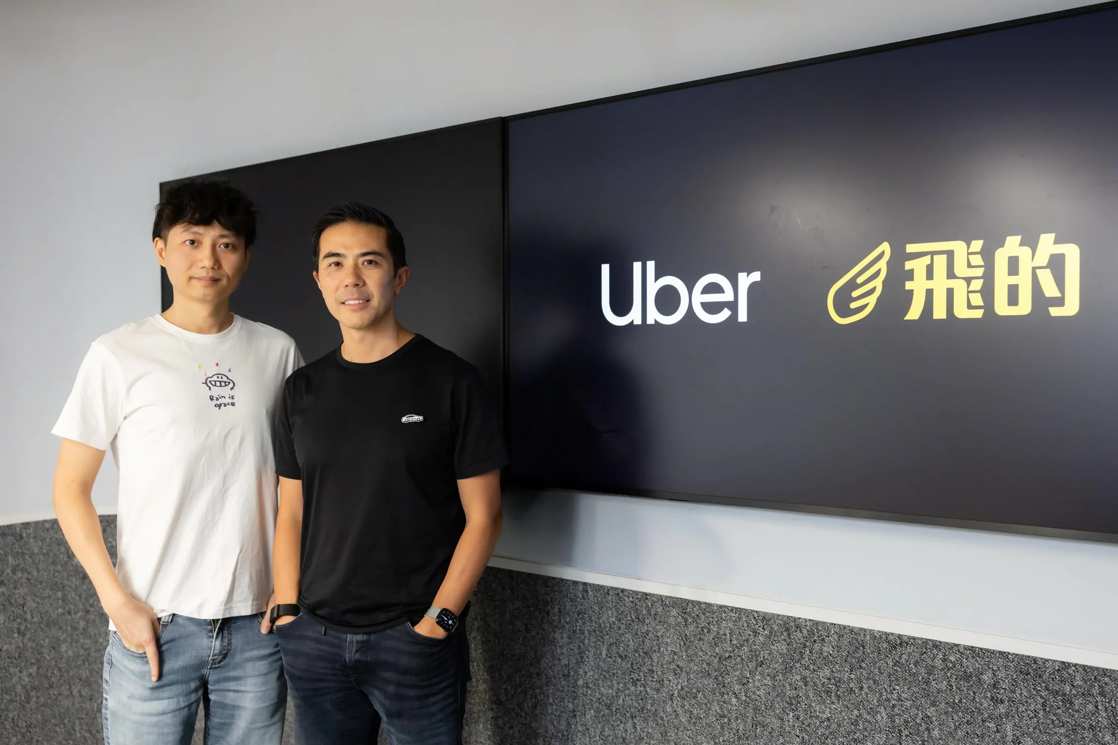 Dois homens em pé em um escritório ao lado de telas exibindo os logotipos da Uber e da empresa Flytaxi