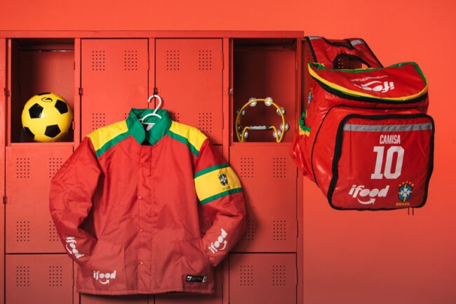 Jaqueta vermelha personalizada do iFood com cores da bandeira do Brasil pendurada em armário esportivo ao lado de mochila térmica, bola de futebol e pandeiro.
