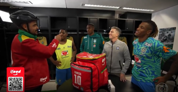 Entregador caracterizado do iFood conversa com jogadores e integrantes da comissão técnica da Seleção Brasileira em vestiário, ao lado de uma bolsa térmica vermelha.