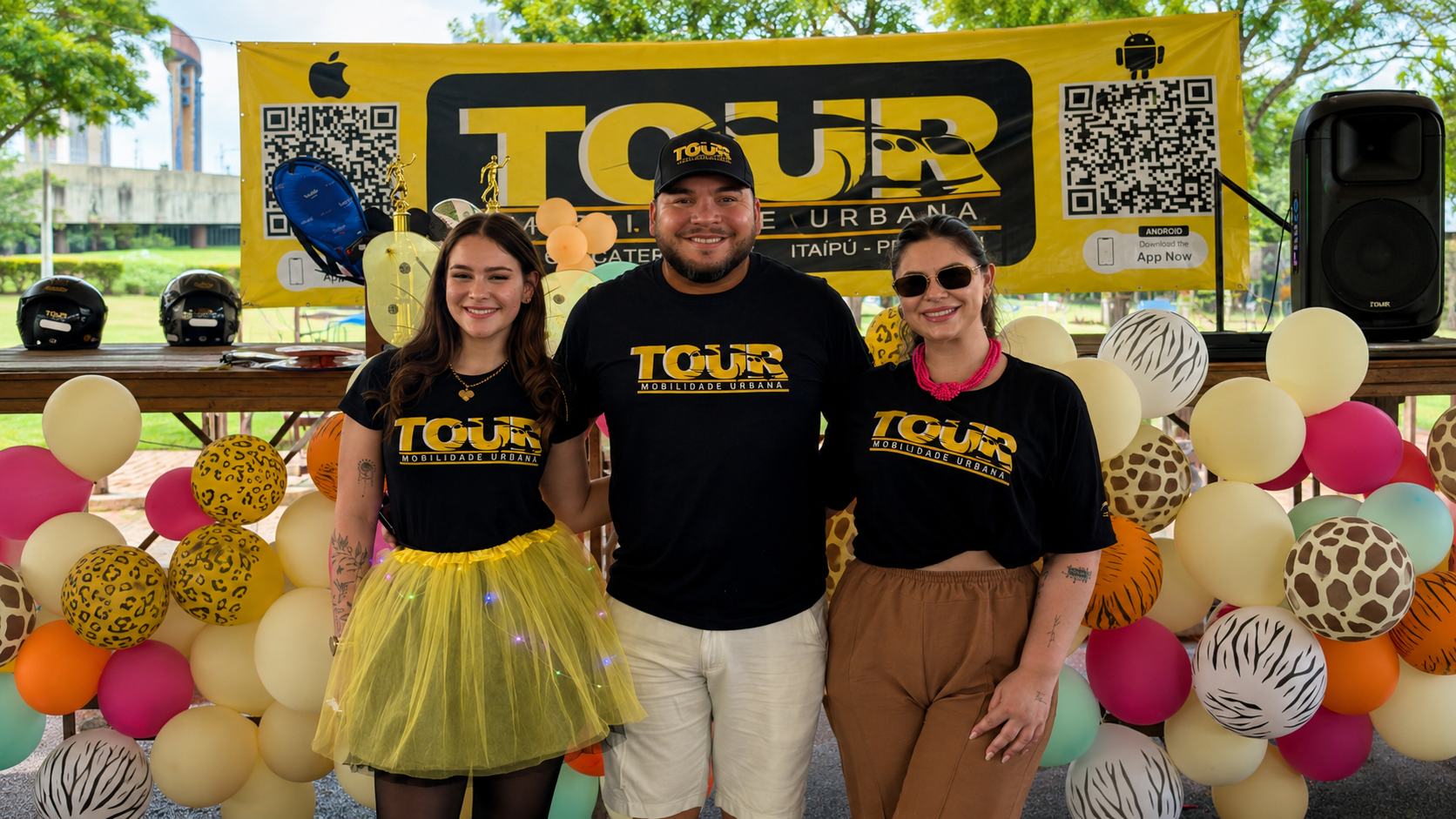 Três pessoas sorrindo em frente a um banner amarelo e preto da Tour Mobilidade Urbana, com balões decorativos, QR codes e itens de exposição ao fundo.
