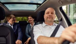 Motorista conduzindo carro executivo com dois passageiros sentados no banco traseiro.