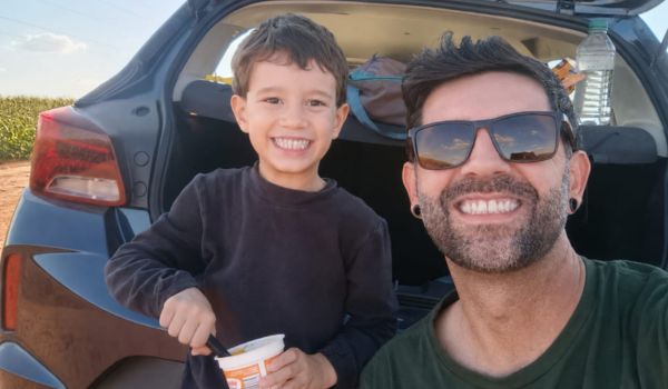 Homem sorrindo usando óculos escuros ao lado de uma criança sorridente em frente ao porta-malas aberto de um carro em área rural.
