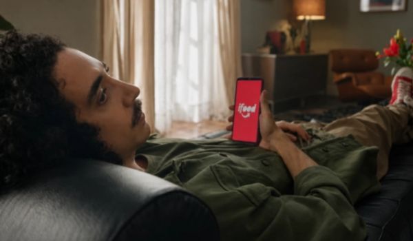 Pessoa deitada em um sofá segurando um smartphone com o aplicativo iFood aberto na tela.