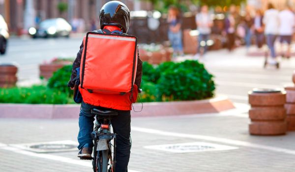 Entregador de delivery em bicicleta usando mochila térmica vermelha em área urbana.