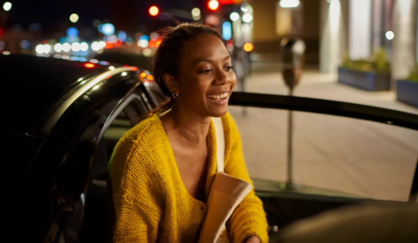 Mulher sorridente usando casaco amarelo entra em um carro à noite, com luzes urbanas desfocadas ao fundo.