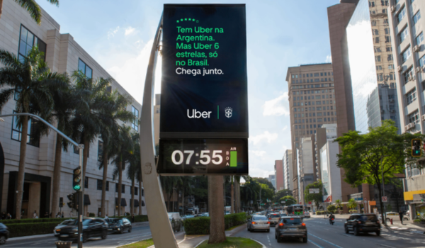 Relógio digital urbano exibe anúncio da Uber sobre a Argentina em uma avenida movimentada, com prédios, carros e árvores ao redor.