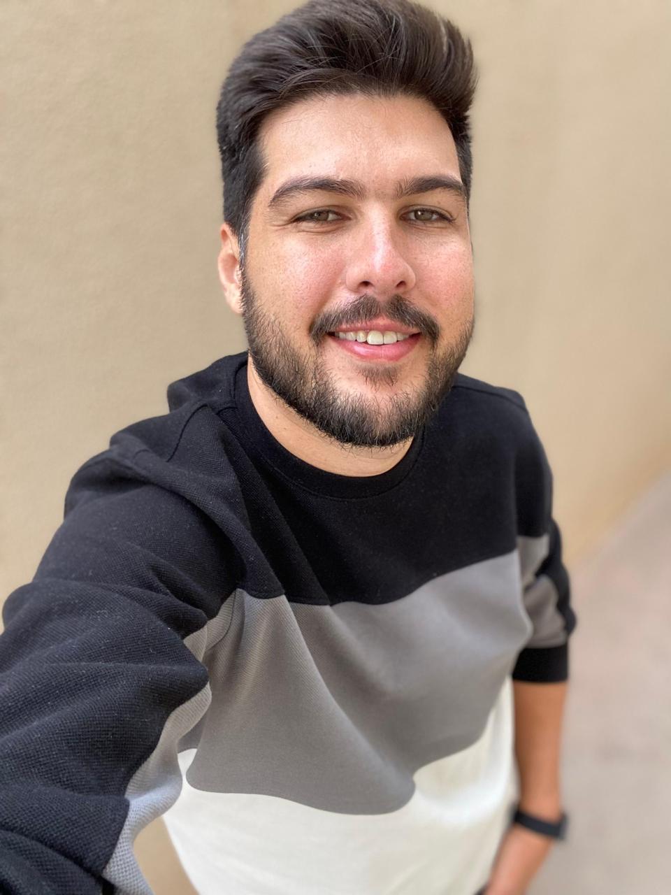 Kelvin Spassini sorrindo para a câmera em um retrato informal, usando blusa preta, cinza e branca, com fundo neutro desfocado.