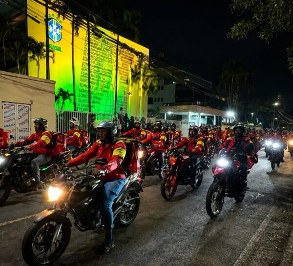 Motociclistas uniformizados em jaquetas vermelhas circulam à noite em frente ao prédio iluminado da Confederação Brasileira de Futebol (CBF).