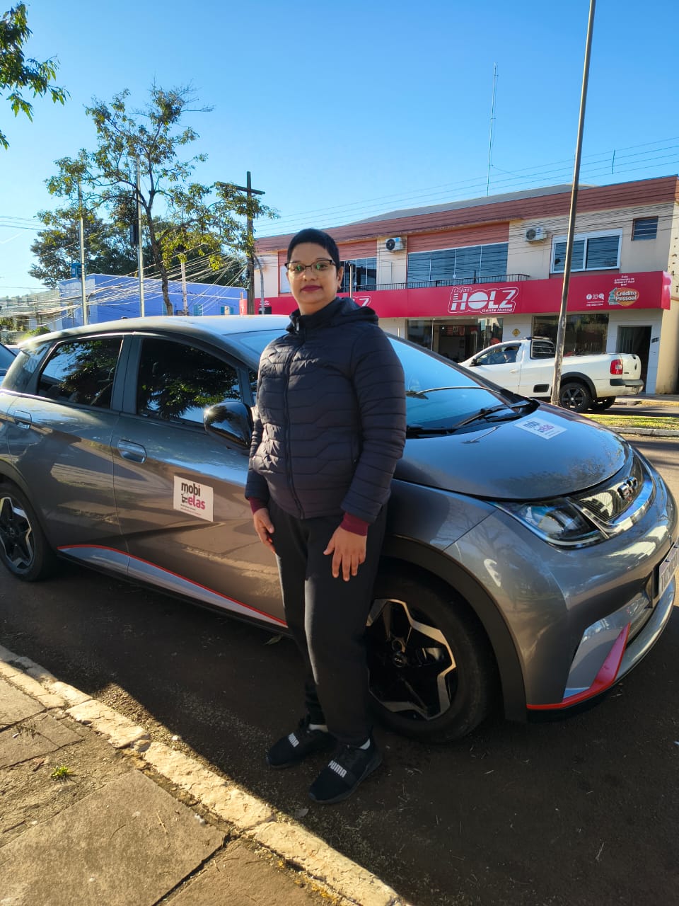 Mulher em pé ao lado de um carro cinza adesivado com a marca Mobi Elas em uma rua comercial durante o dia.