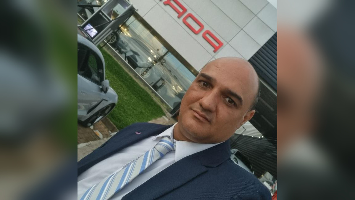 Homem de terno e gravata em selfie diante de uma fachada comercial com carros ao fundo.