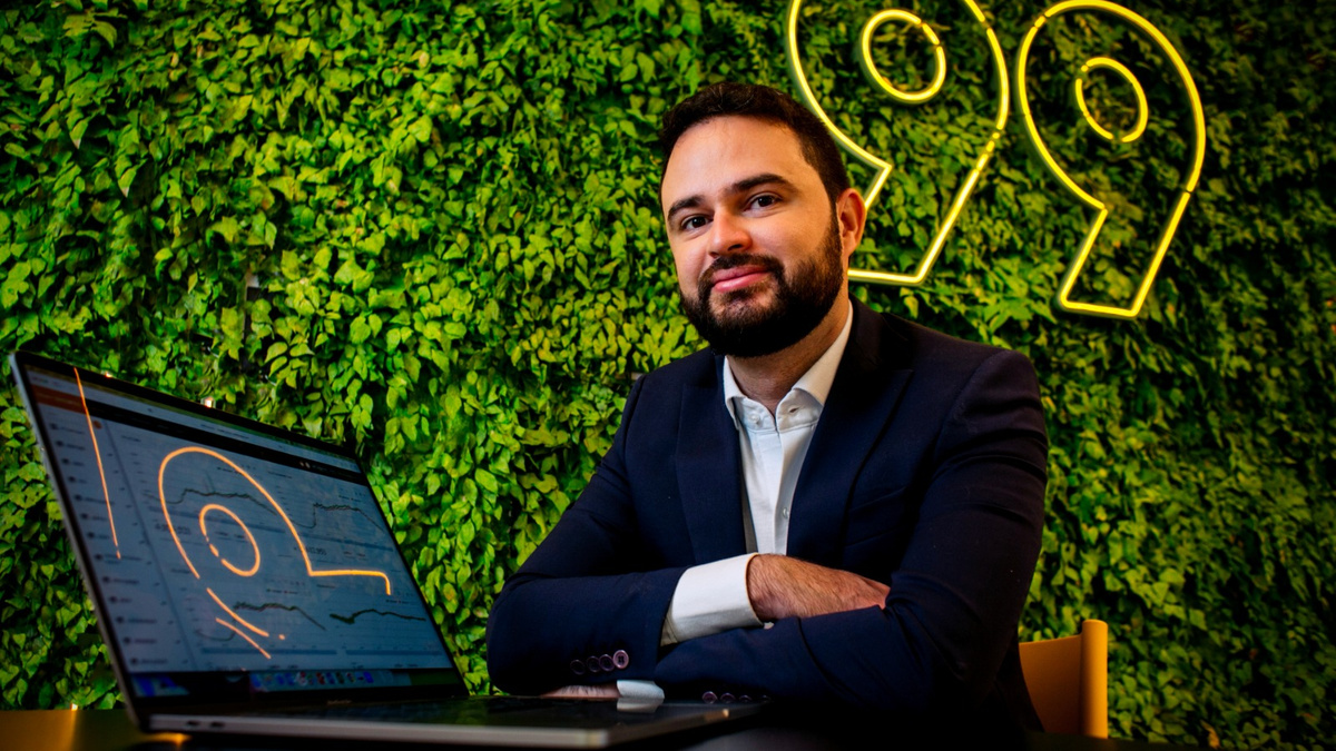 Homem de terno sentado à mesa com um notebook, diante de uma parede verde com letreiro luminoso ao fundo.