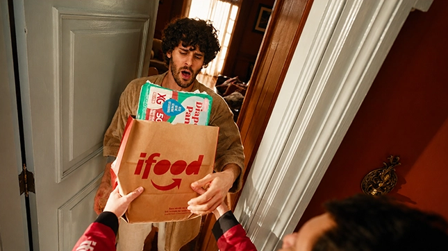 Entregador do iFood entrega sacola com fraldas descartáveis e produtos infantis na porta de uma residência.