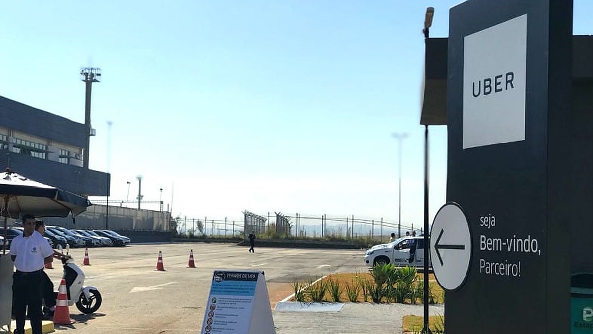 Placa da Uber ao lado de estacionamento com veículos, cones de sinalização e uma pessoa em área externa.