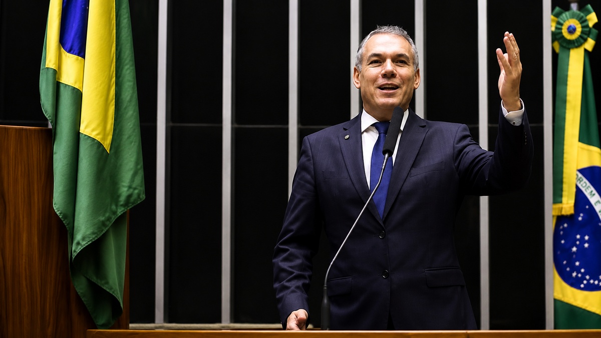 Homem de terno escuro discursa diante de um microfone, com a mão direita levantada e bandeiras do Brasil ao fundo.