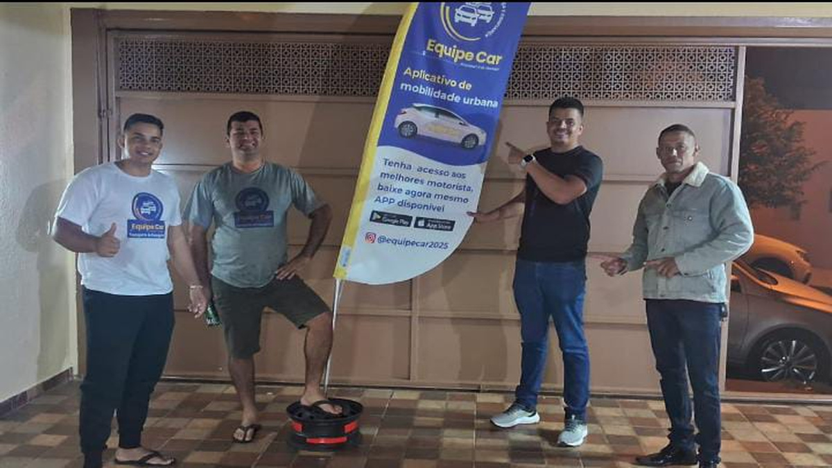 Quatro homens posam em uma área coberta ao lado de uma faixa vertical do aplicativo Equipe Car, com um automóvel parcialmente visível ao fundo.