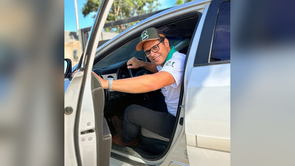 Homem de boné e óculos sorri sentado ao volante de um veículo branco com a porta aberta.