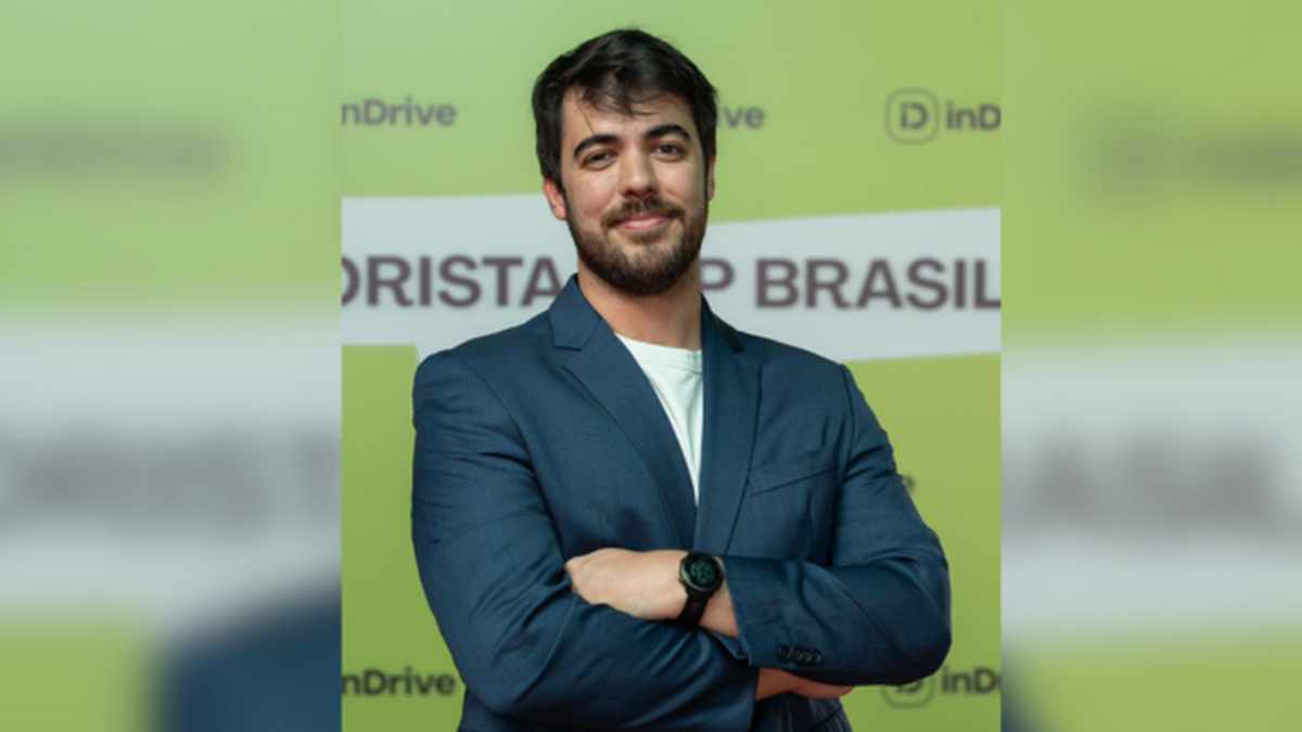 Homem de barba, usando blazer azul e camiseta branca, posa de braços cruzados diante de um painel verde com a marca inDrive.