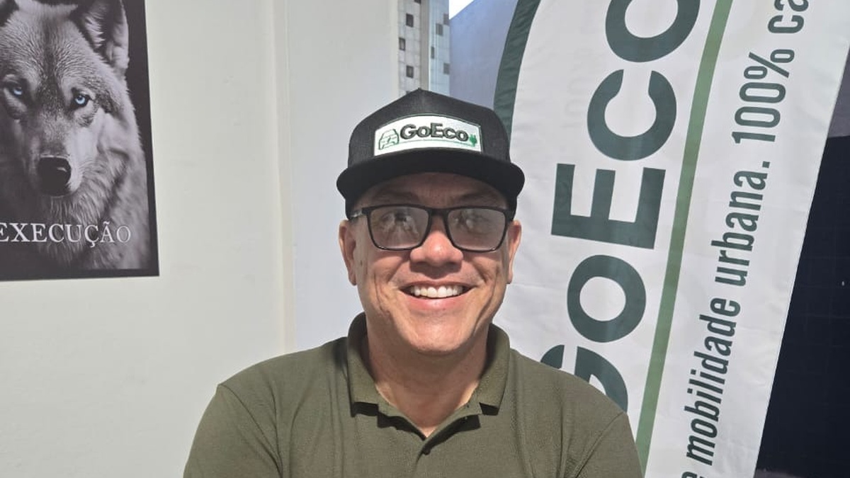 Homem sorridente com boné, óculos e camisa da GoEco, de braços cruzados diante de um banner da empresa.