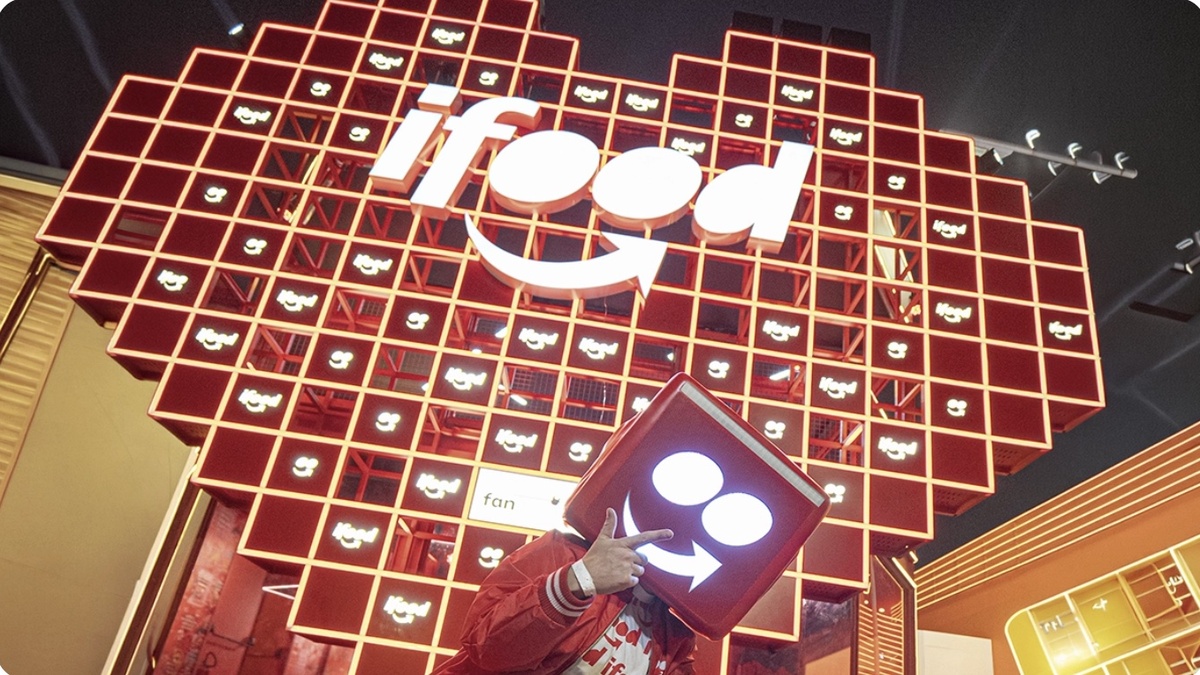 Pessoa com jaqueta vermelha e adereço cúbico na cabeça aponta para o símbolo do iFood diante de uma estrutura iluminada da marca.