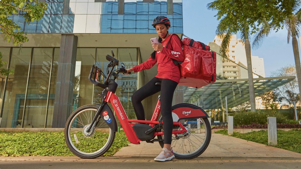 Pessoa com uniforme vermelho e mochila de entrega consulta o celular ao lado de uma bicicleta elétrica diante de um edifício.