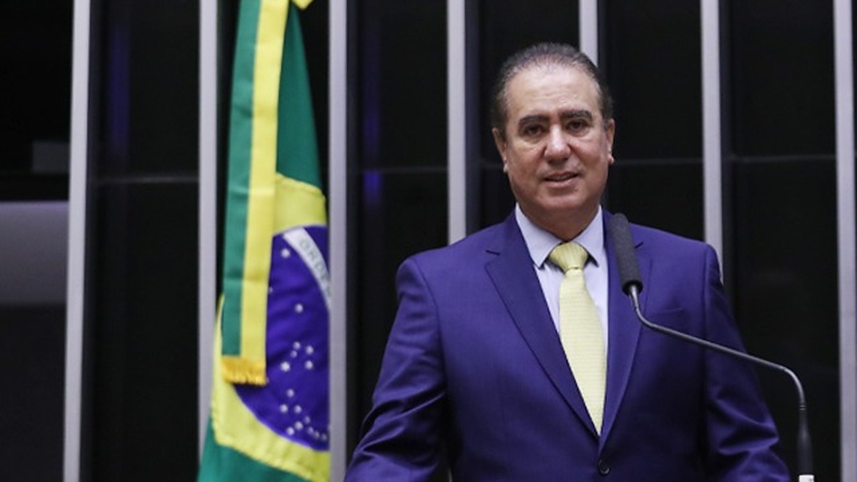Homem de terno discursa diante de um microfone, com a bandeira do Brasil ao fundo.
