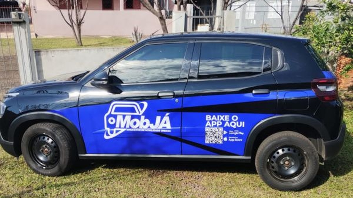 Carro preto e azul com adesivo da MobJá estacionado em área gramada.