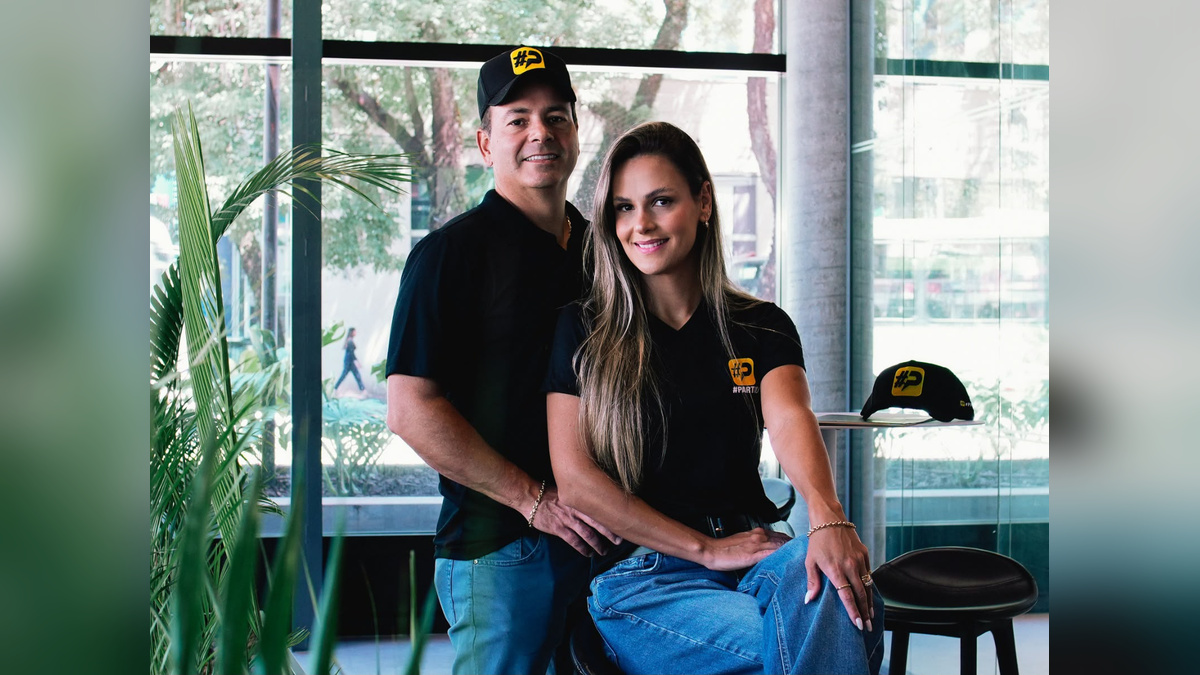 Homem e mulher sorrindo usam camisetas pretas com símbolo amarelo e posam em ambiente interno com grandes janelas e plantas.