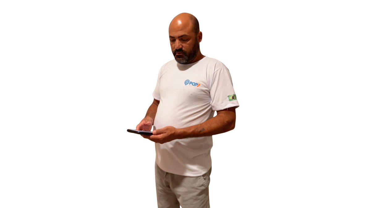 Homem de barba usa camiseta branca com a marca POP7 enquanto manuseia um celular.