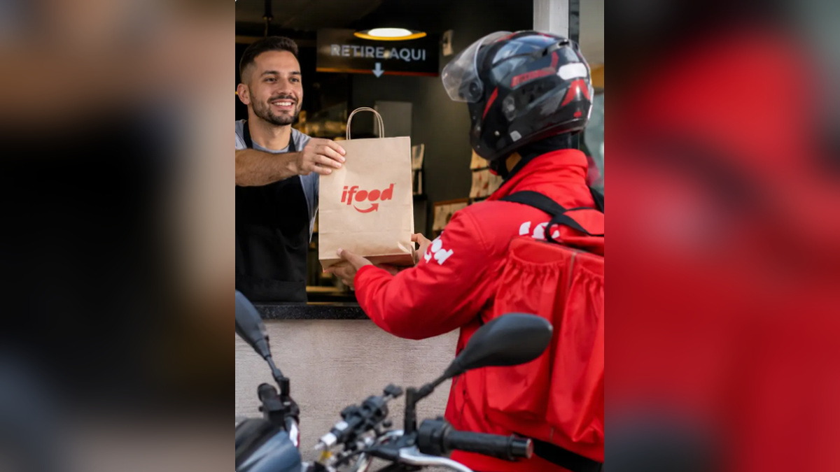 Atendente entrega uma sacola do iFood a um entregador de capacete e mochila vermelha em um balcão de retirada.