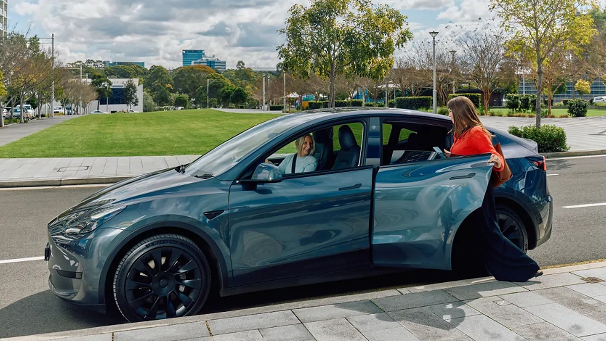 Mulher abre a porta traseira de um carro azul estacionado enquanto outra mulher permanece no banco do motorista.