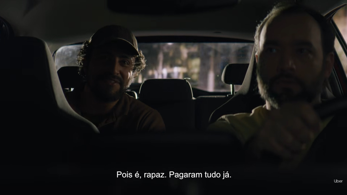 Dois homens aparecem no interior de um carro durante a noite, com uma legenda na parte inferior da cena.