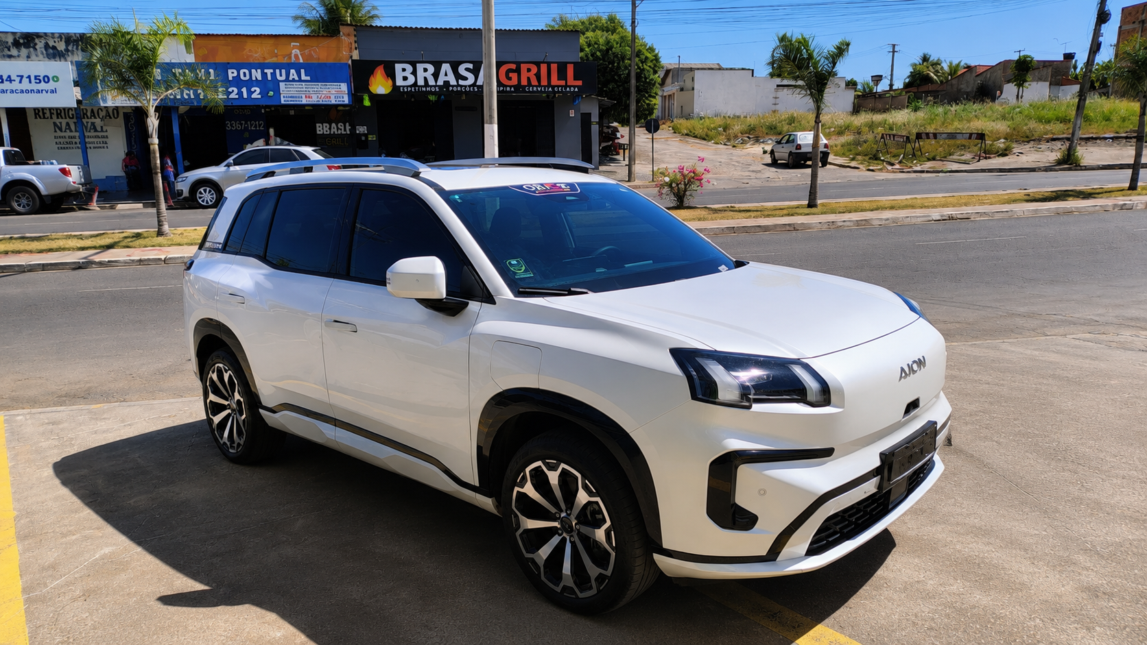 SUV branco estacionado em área externa, visto de frente e de lado, com estabelecimentos comerciais e uma avenida ao fundo.