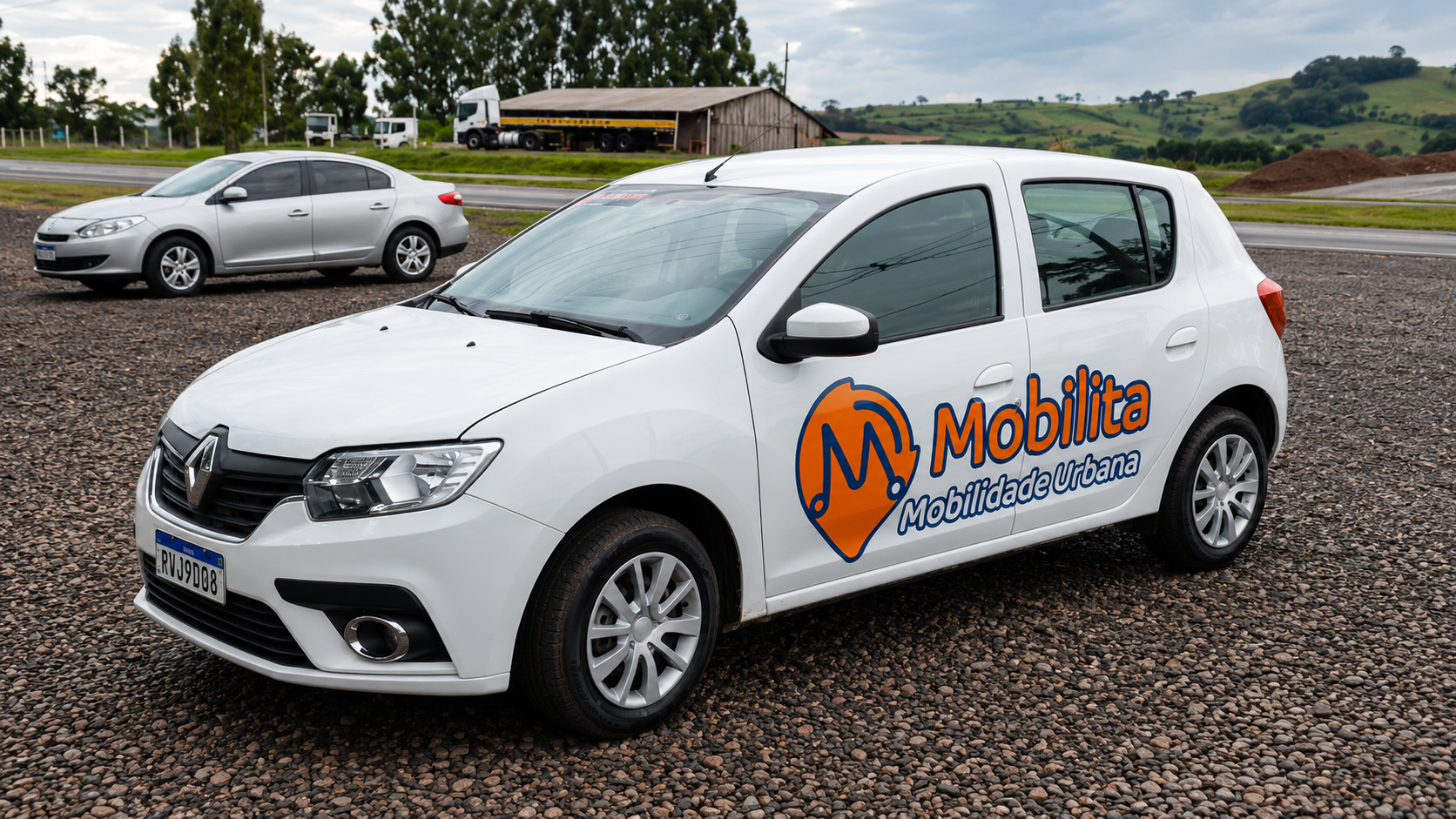Carro branco da Mobilita Mobilidade Urbana estacionado em um terreno de pedras, próximo a uma rodovia. O veículo possui o logotipo laranja e azul da empresa nas portas laterais.