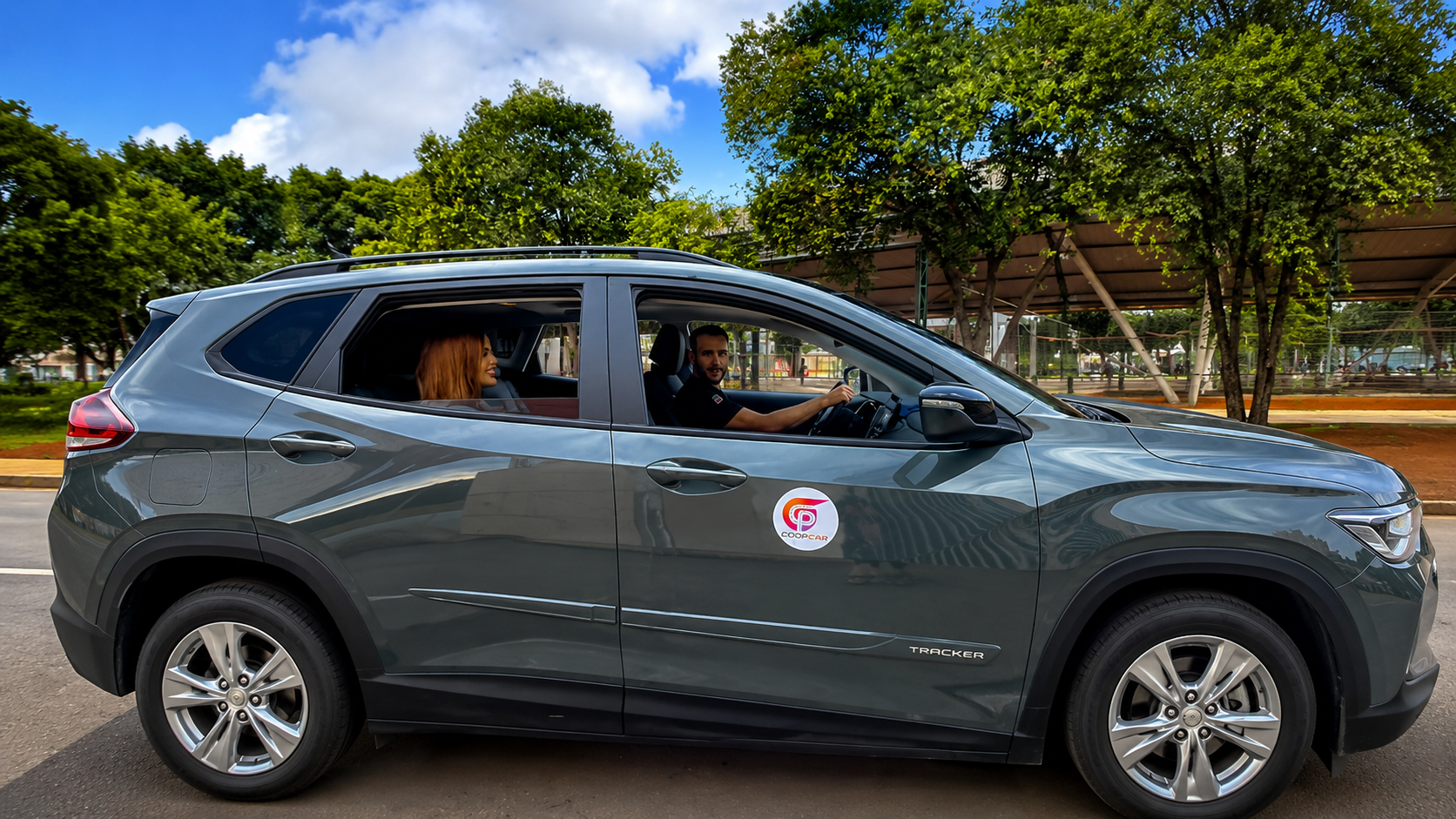 Carro cinza com adesivo da Coopcar na lateral, com um homem ao volante e uma mulher no banco traseiro, em área arborizada.