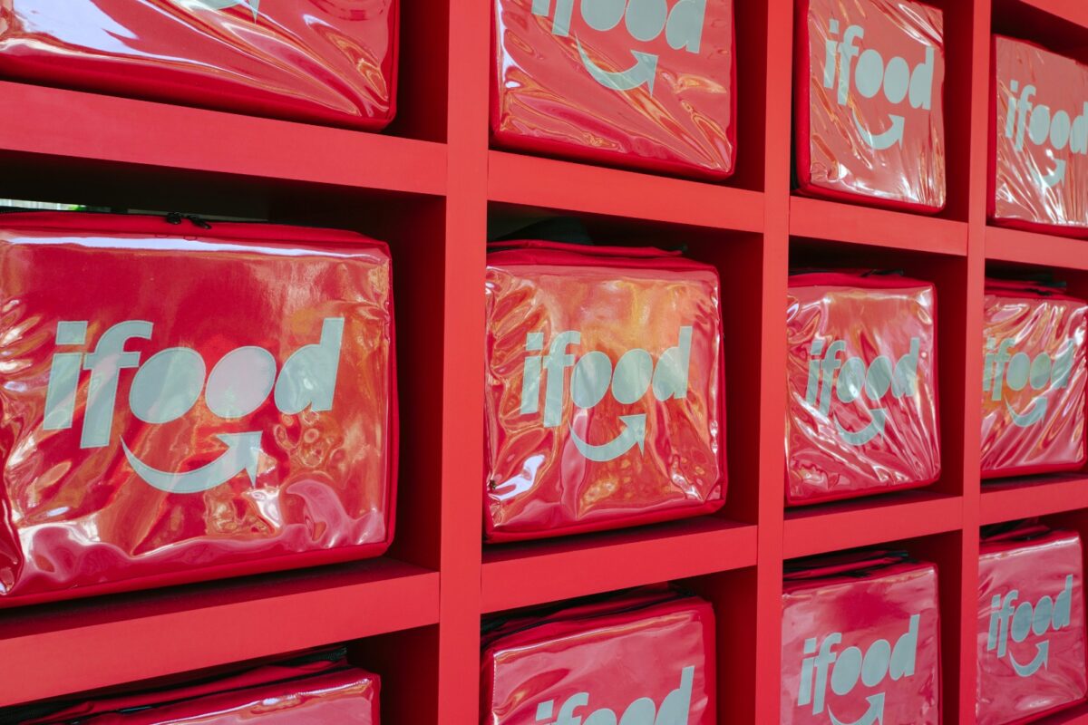 Bolsas térmicas vermelhas de entrega com a marca iFood são exibidas em prateleiras da mesma cor.