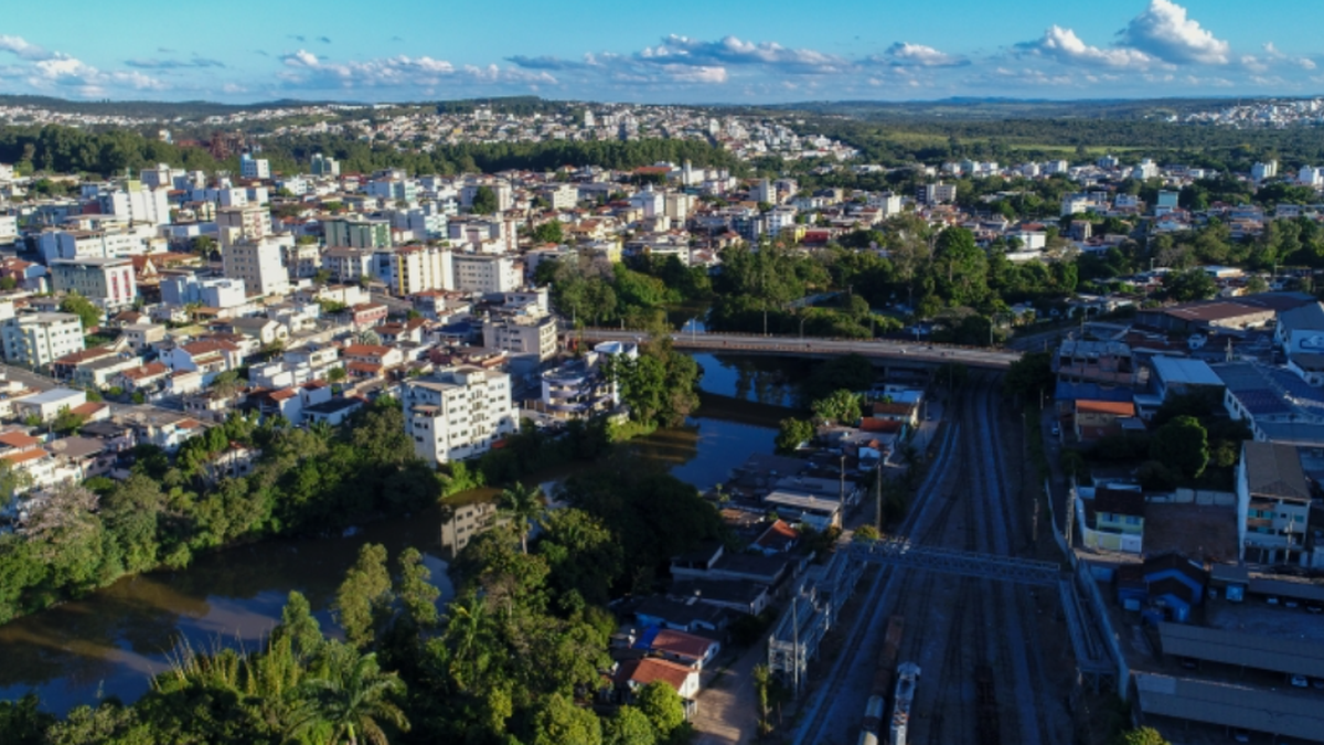 Vista aérea da cidade de Divinópolis (MG), com prédios, casas, áreas verdes, rio e linha férrea.